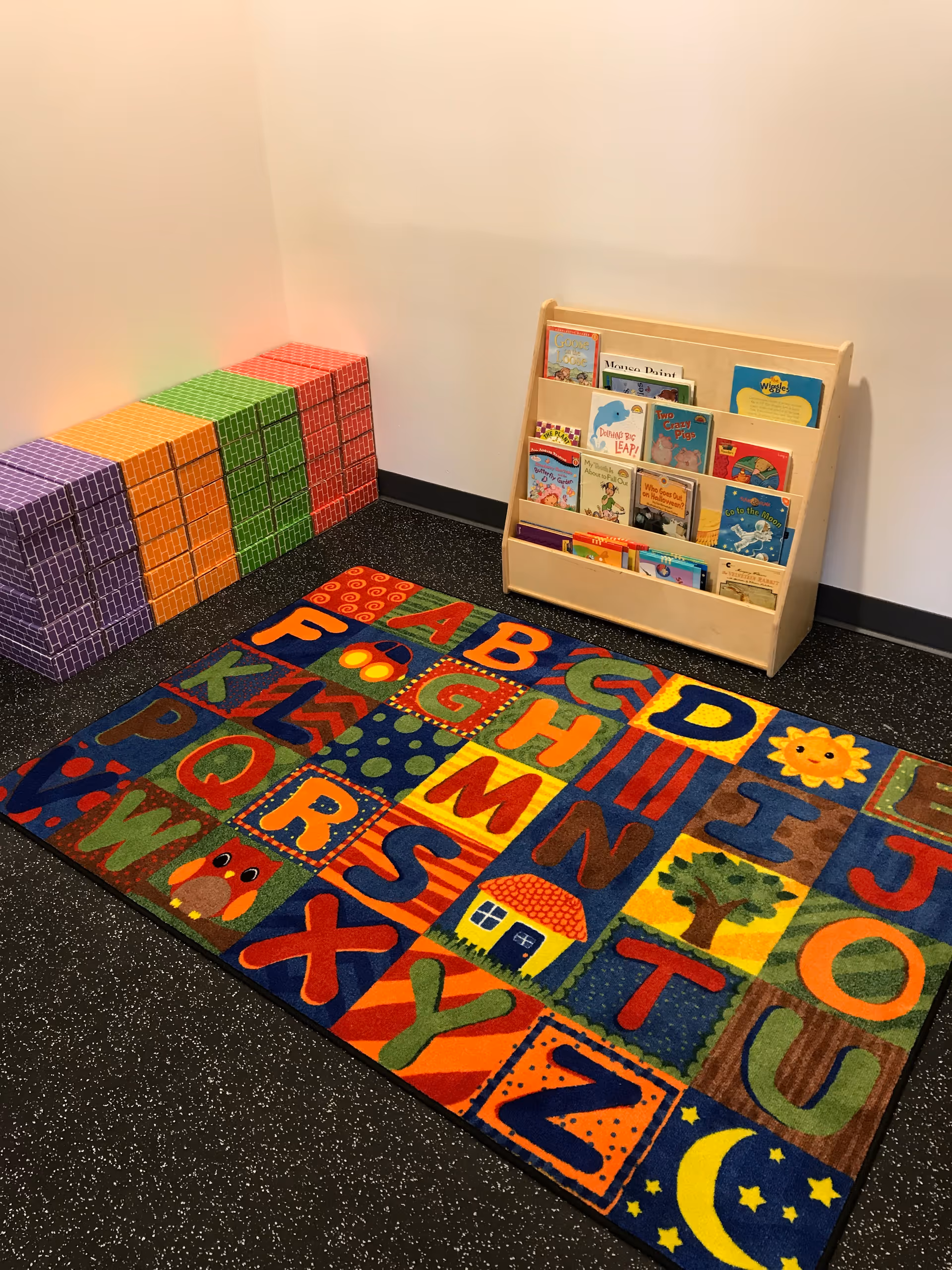 Bright interior corner with a colorful alphabet rug, stacked blocks, and a wooden book rack.