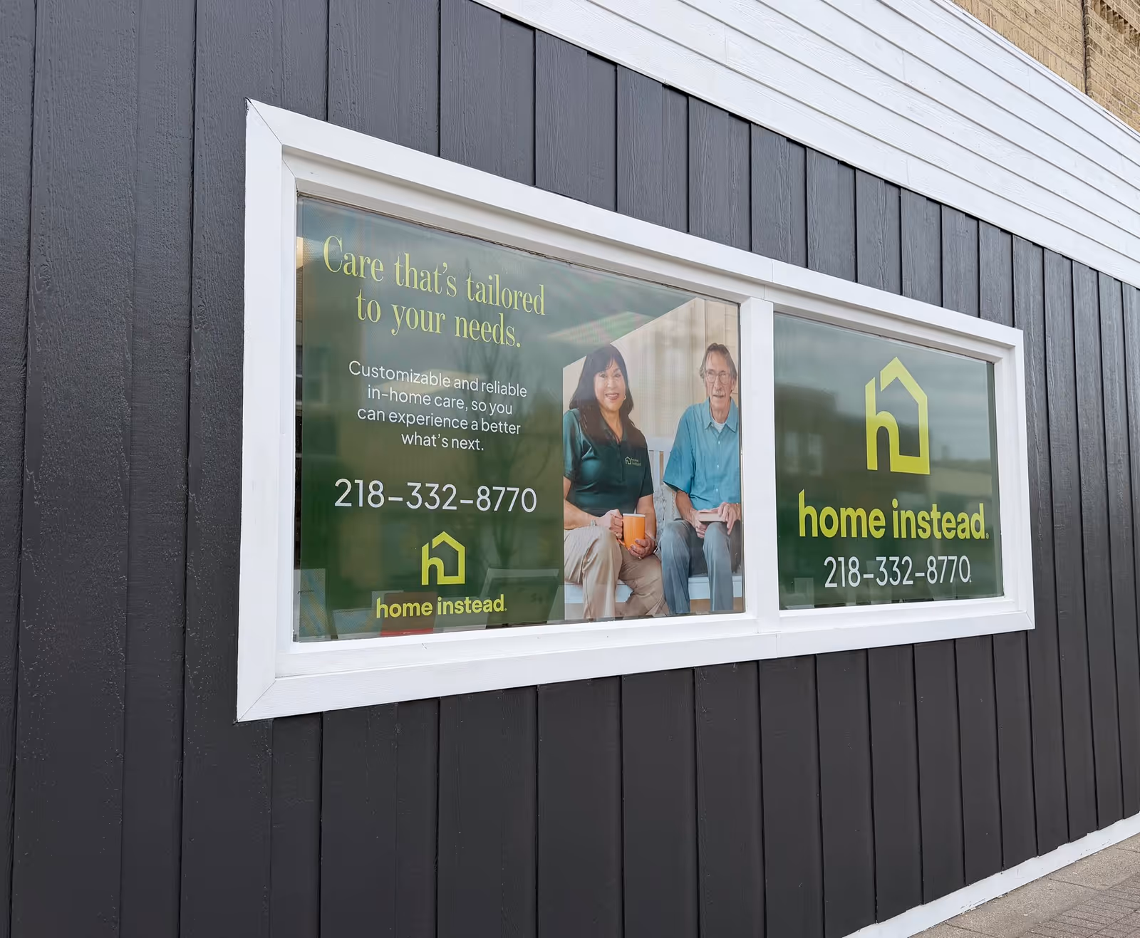 Window advertisement on a dark wooden exterior wall featuring two people and Home Instead signage.