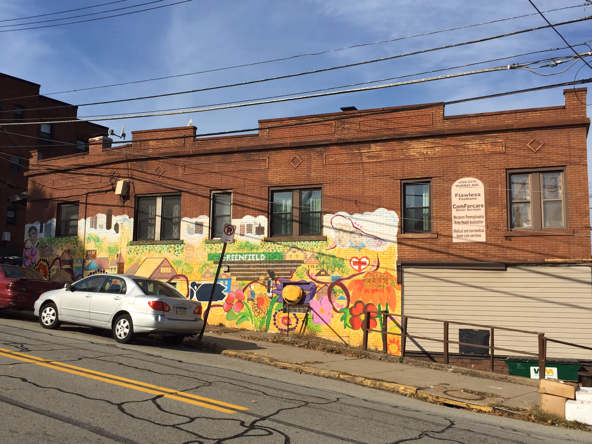 Brick building with a colorful mural along the wall and parked cars on the street.