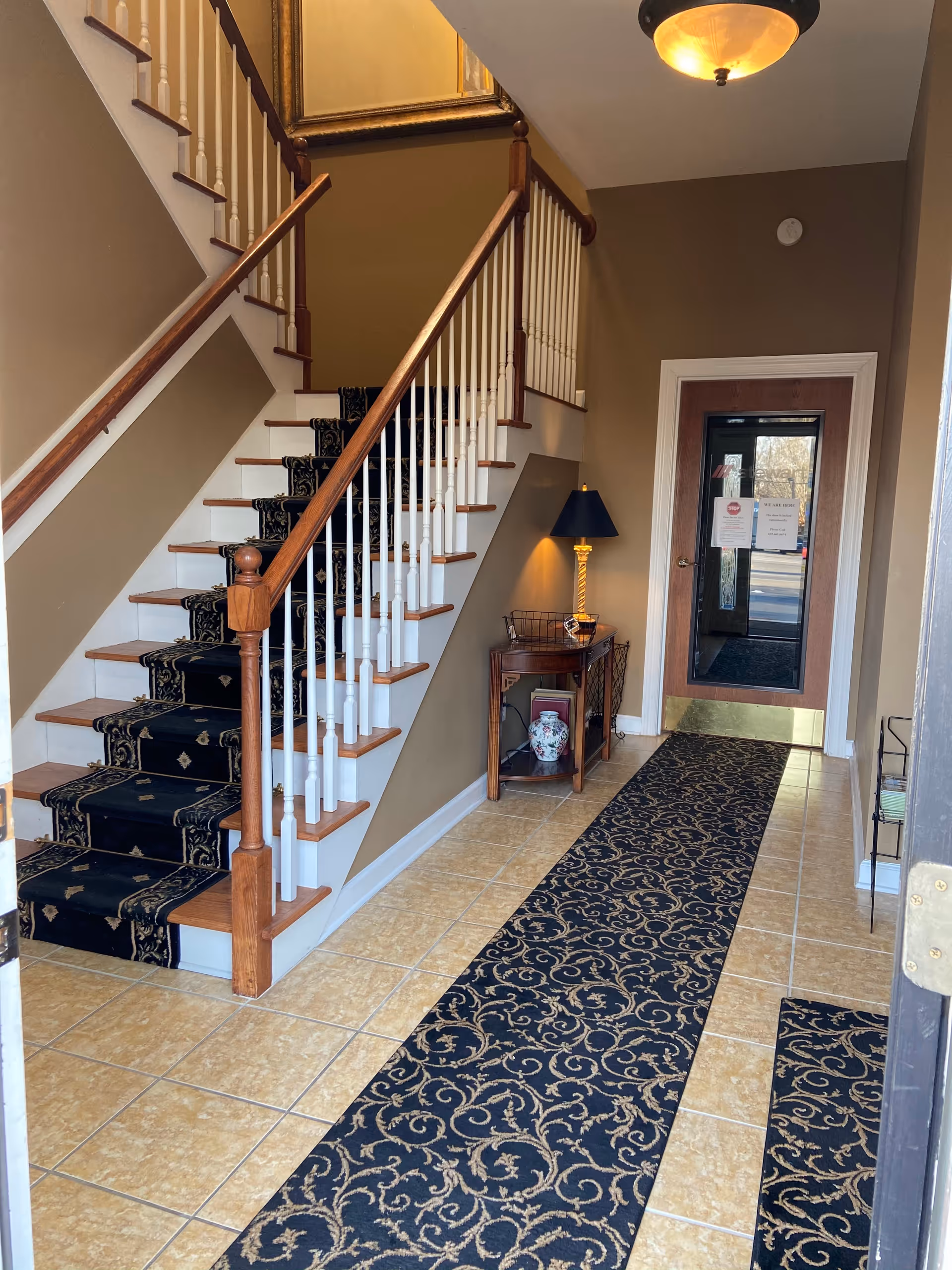 Interior foyer with carpeted staircase, decorative railing, side table, lamp, and entry door.