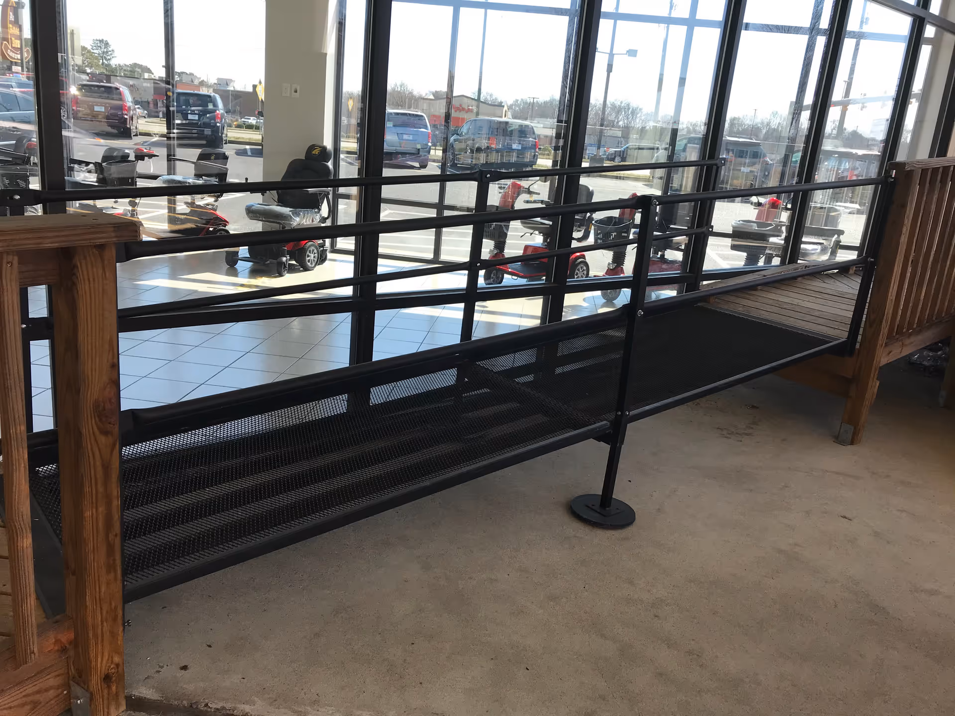 Indoor ramp with black metal railing beside glass storefront windows overlooking a parking lot.