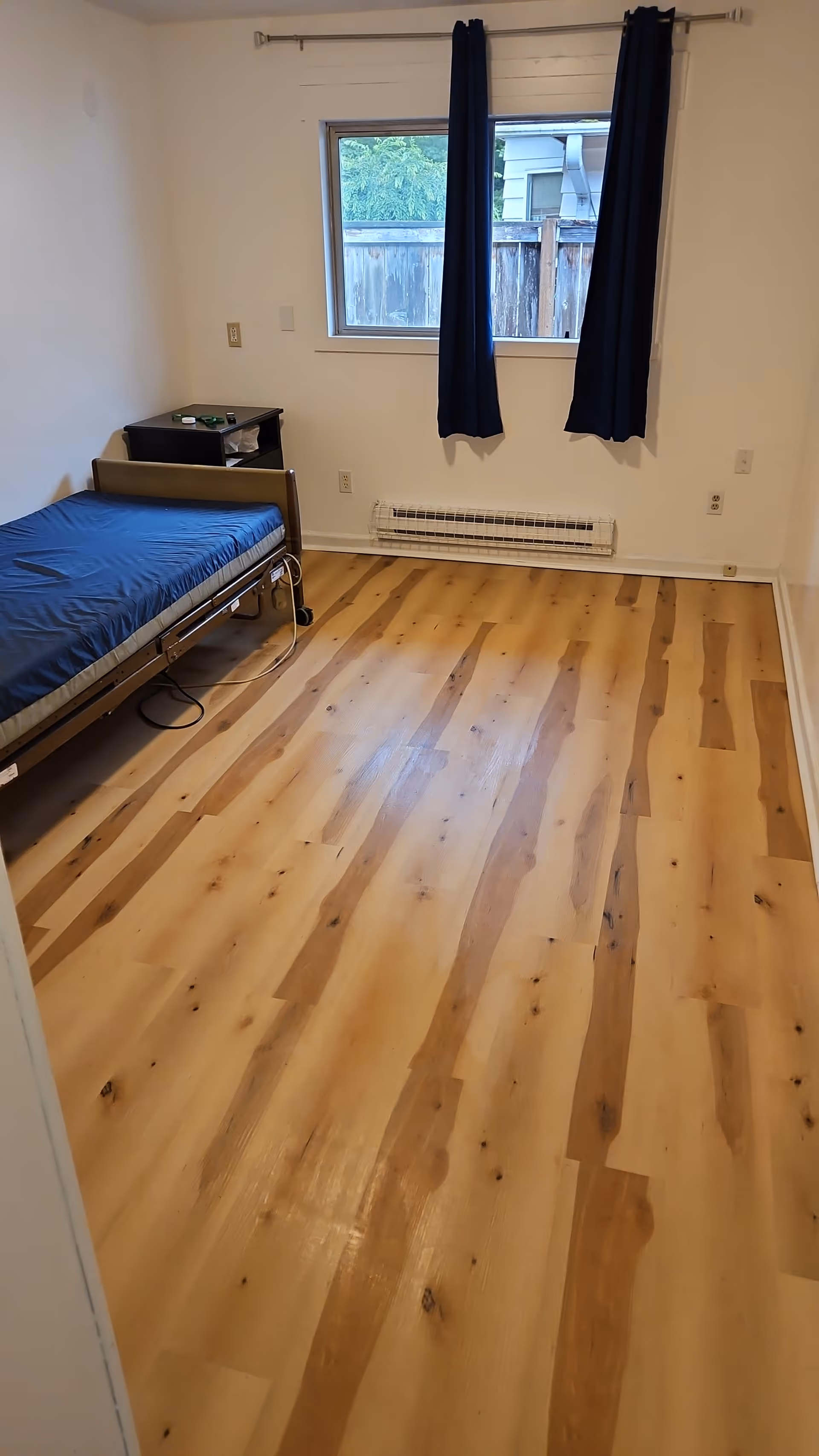 Small bedroom with single bed, nightstand, window, blue curtains, and wooden floor.