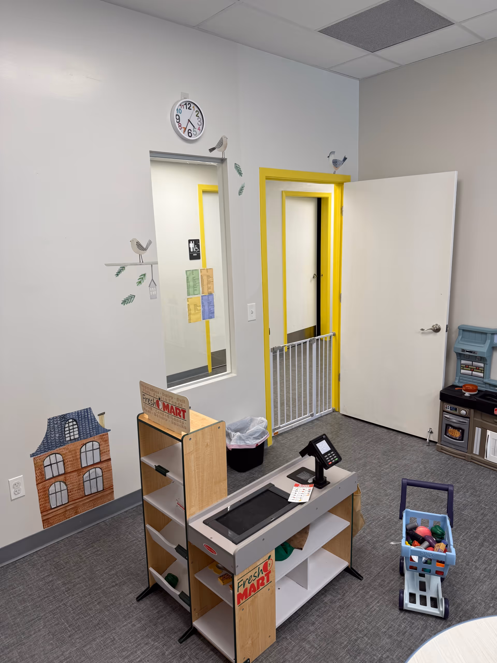 Interior therapy play area with checkout counter, toy kitchen, shelves, and wall murals.