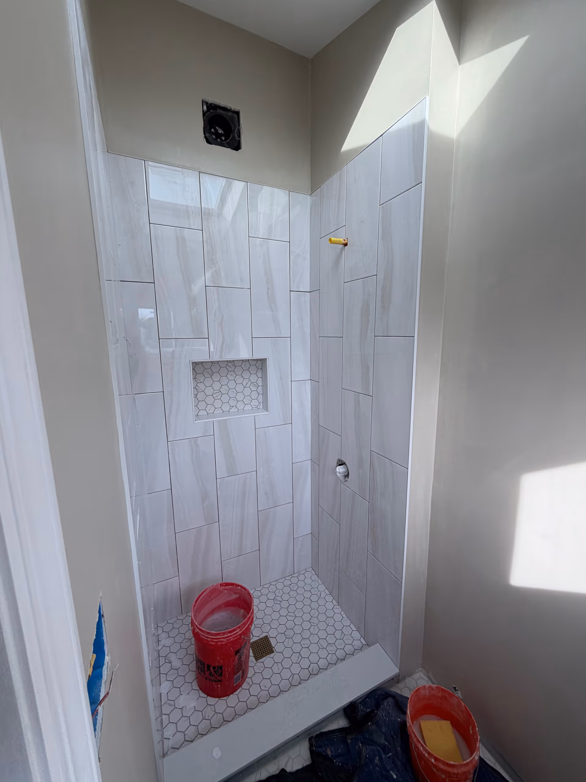 Unfinished bathroom shower with white tiles, built in niche, and red buckets on floor.
