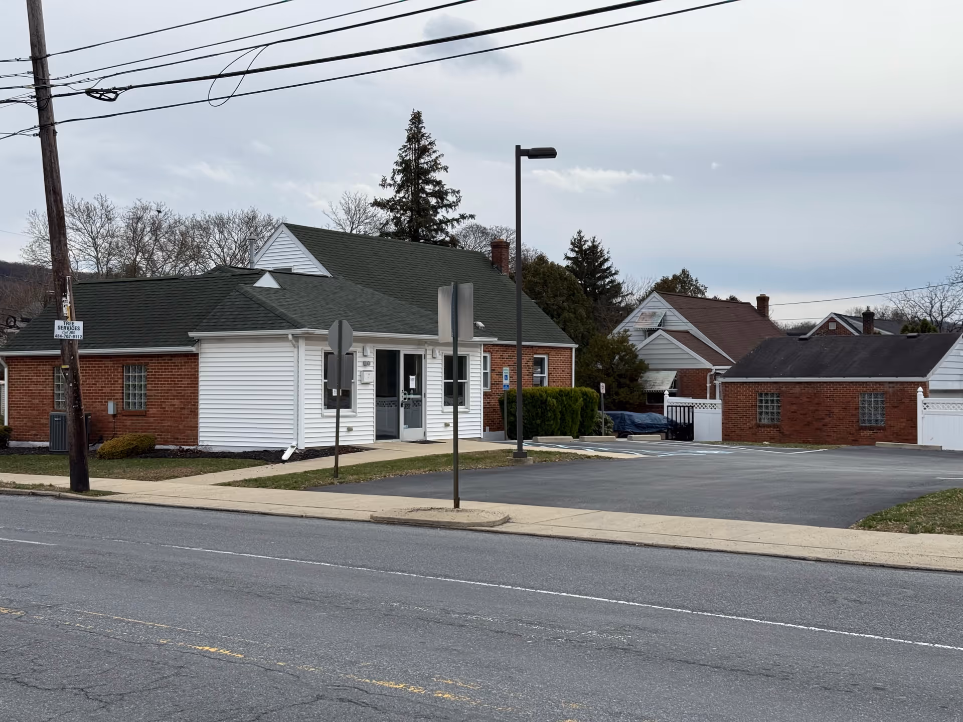 Front view of a small brick building with white siding, parking lot, and a sidewalk.