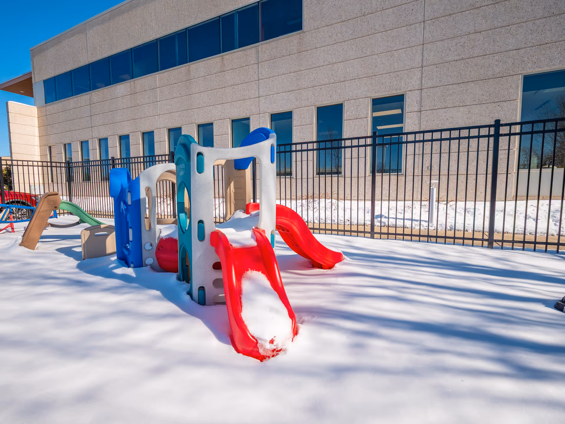 Snow-covered playground with colorful slides beside a beige building and black fence.