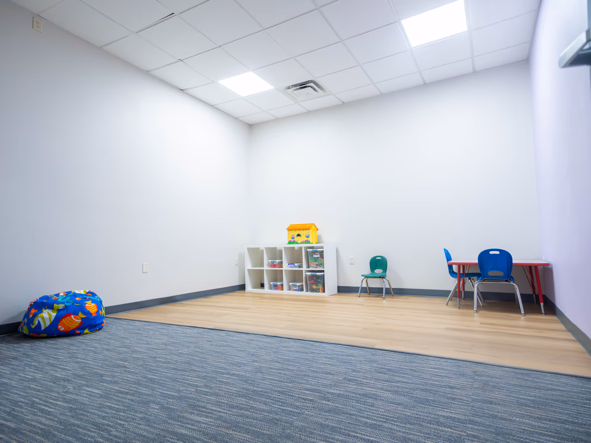 An empty children's playroom with toy shelves, bins, bean bag, and small tables.