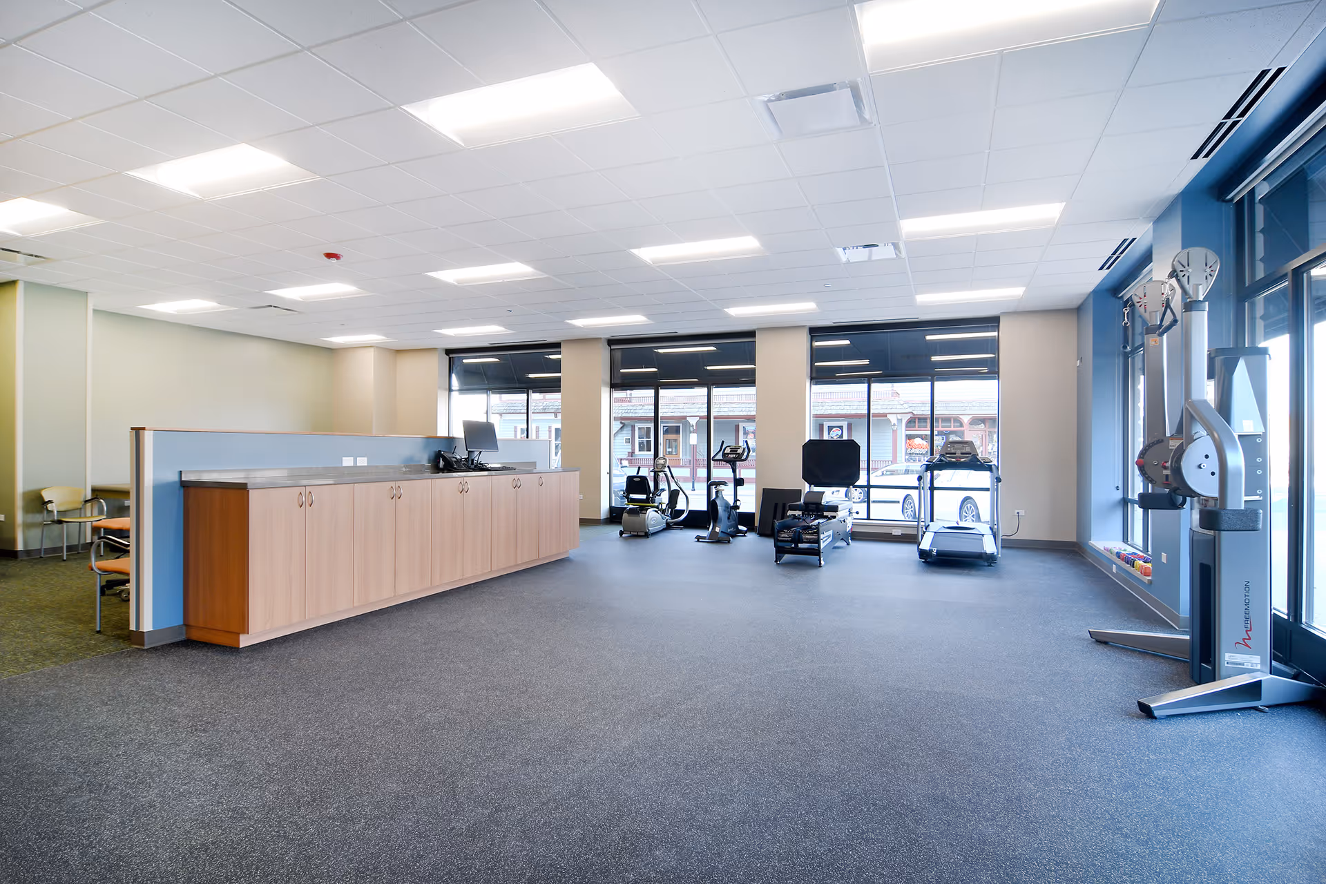 A spacious physical therapy gym with cardio machines, a front desk, and large windows.