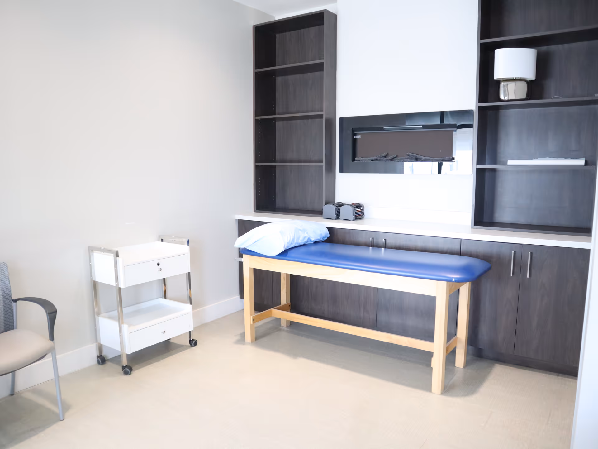 A treatment table with blue cushion in a clean room with dark wood cabinets and shelves.