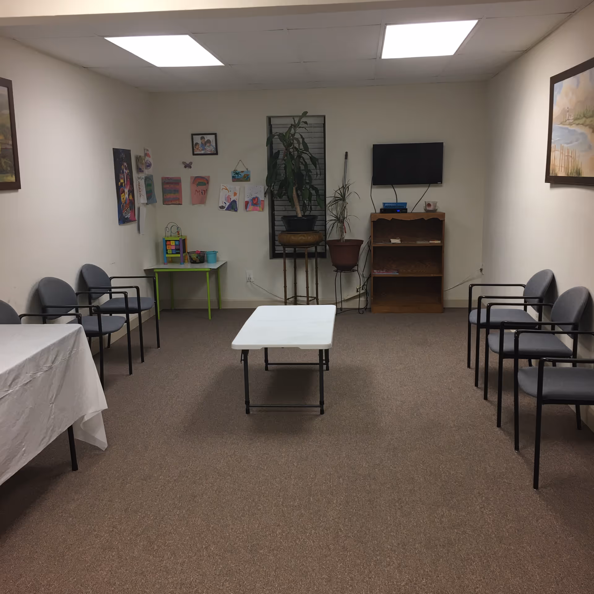 A small common room with chairs lined along walls, a central folding table, and a TV.