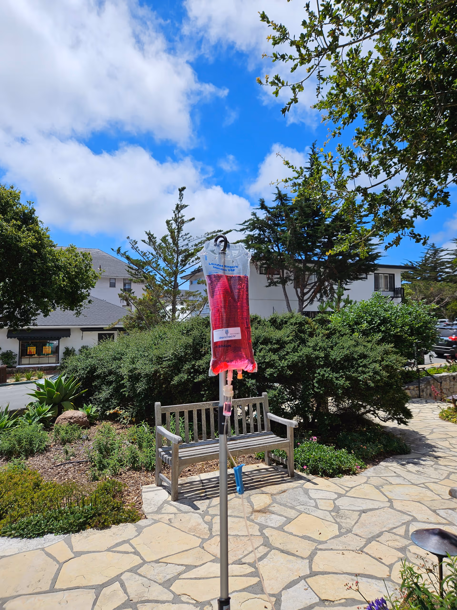 Outdoor courtyard with a bench and pink IV bag on a pole under a blue sky.