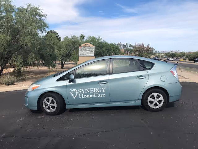 Light blue Synergy HomeCare vehicle parked in a lot with facility sign and trees behind.