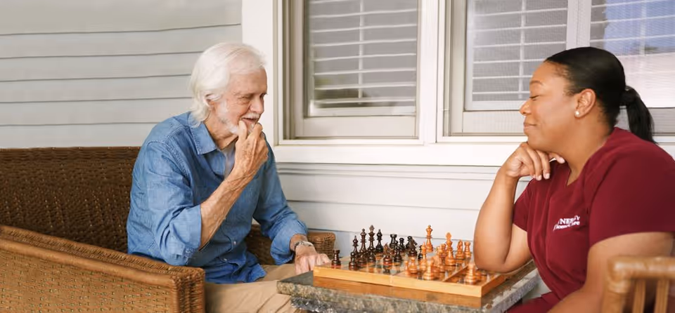 Elderly man and caregiver sit on a porch, playing chess together.