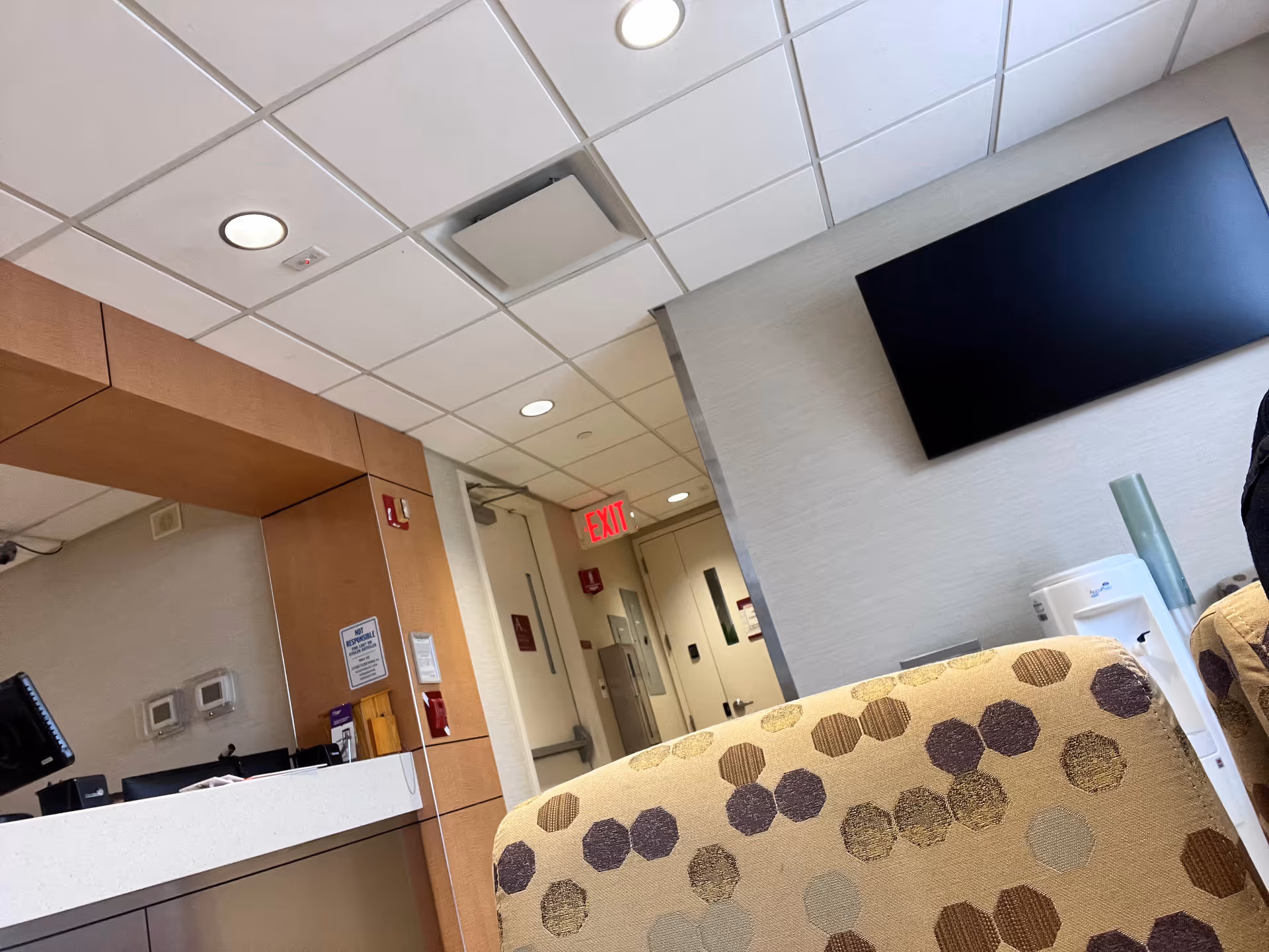 Inside a senior living facility lobby with a patterned chair, reception desk, and wall-mounted TV.