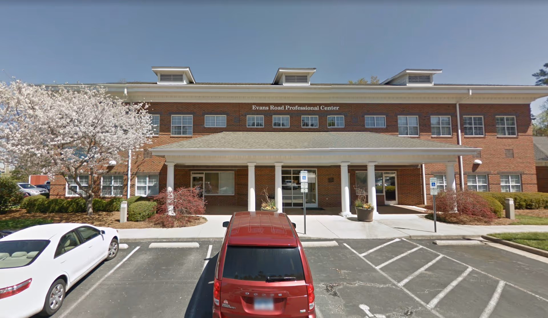 Brick building with a front entrance and columns, parking lot with several cars.