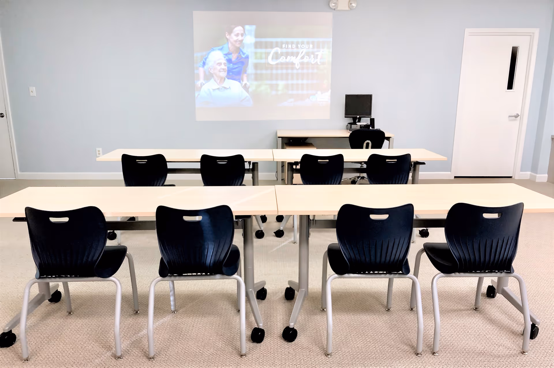 Bright interior classroom with rows of light desks, dark chairs, and a projector screen.