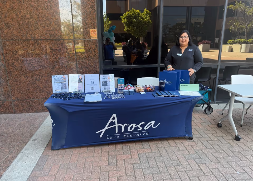 A staff member stands behind a blue Arosa promotional table outside a building entrance.