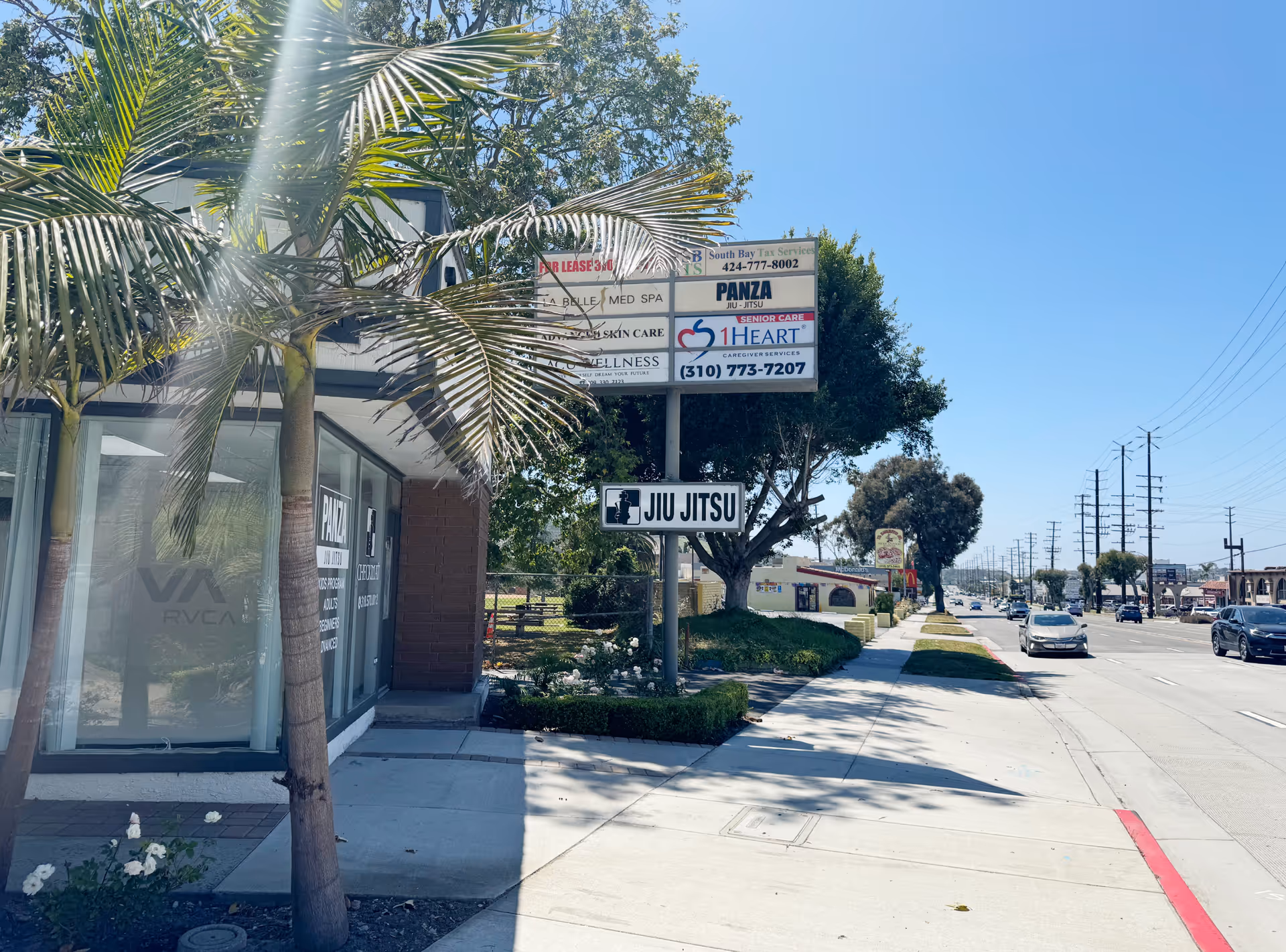 Exterior street view of a building with 1Heart Caregiver Services sign and palm trees lining sidewalk