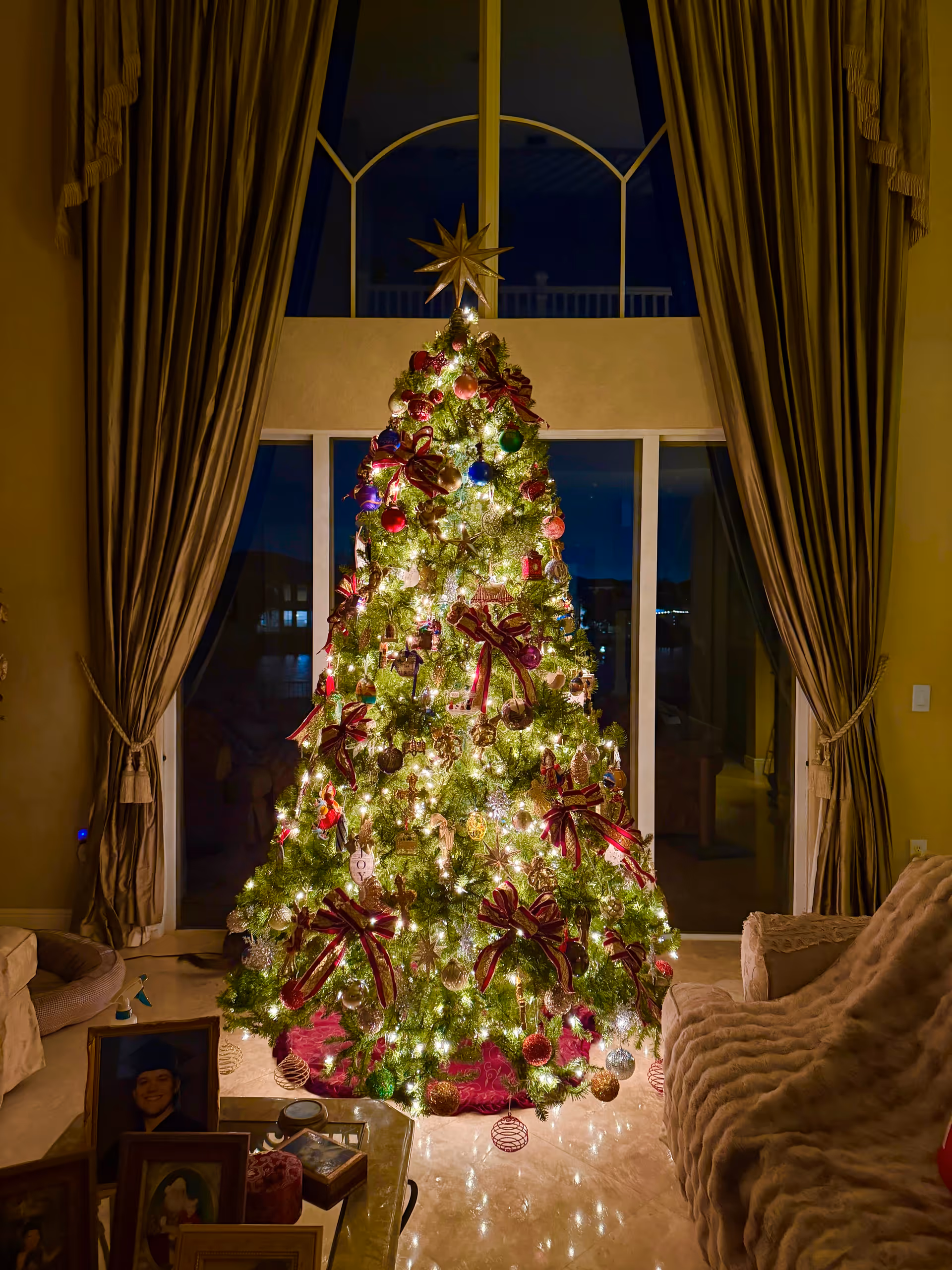 A lit Christmas tree stands in a living room with curtains and family photos nearby.