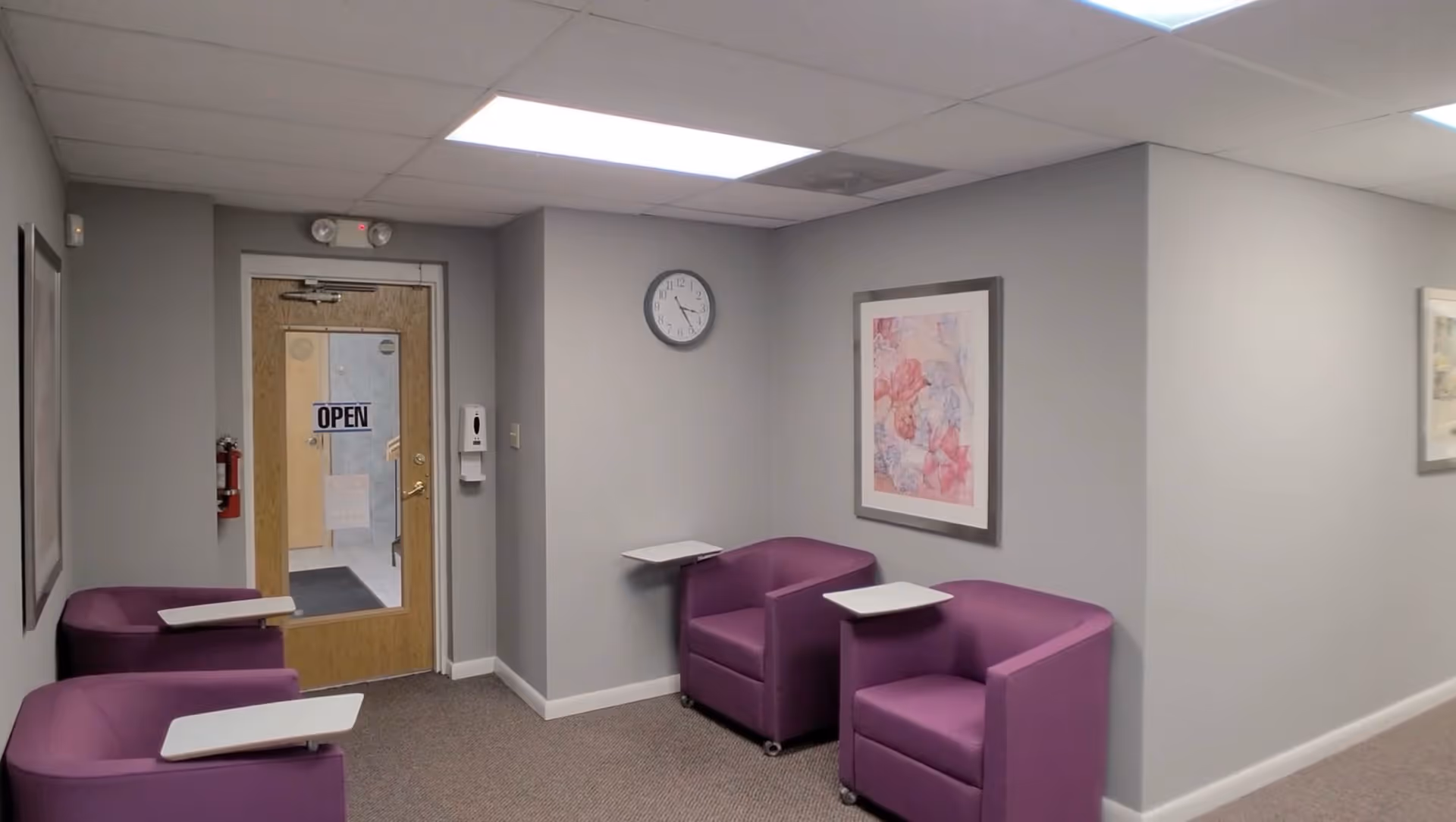Interior lounge area with purple chairs, small desks, framed art, clock, and doorway.