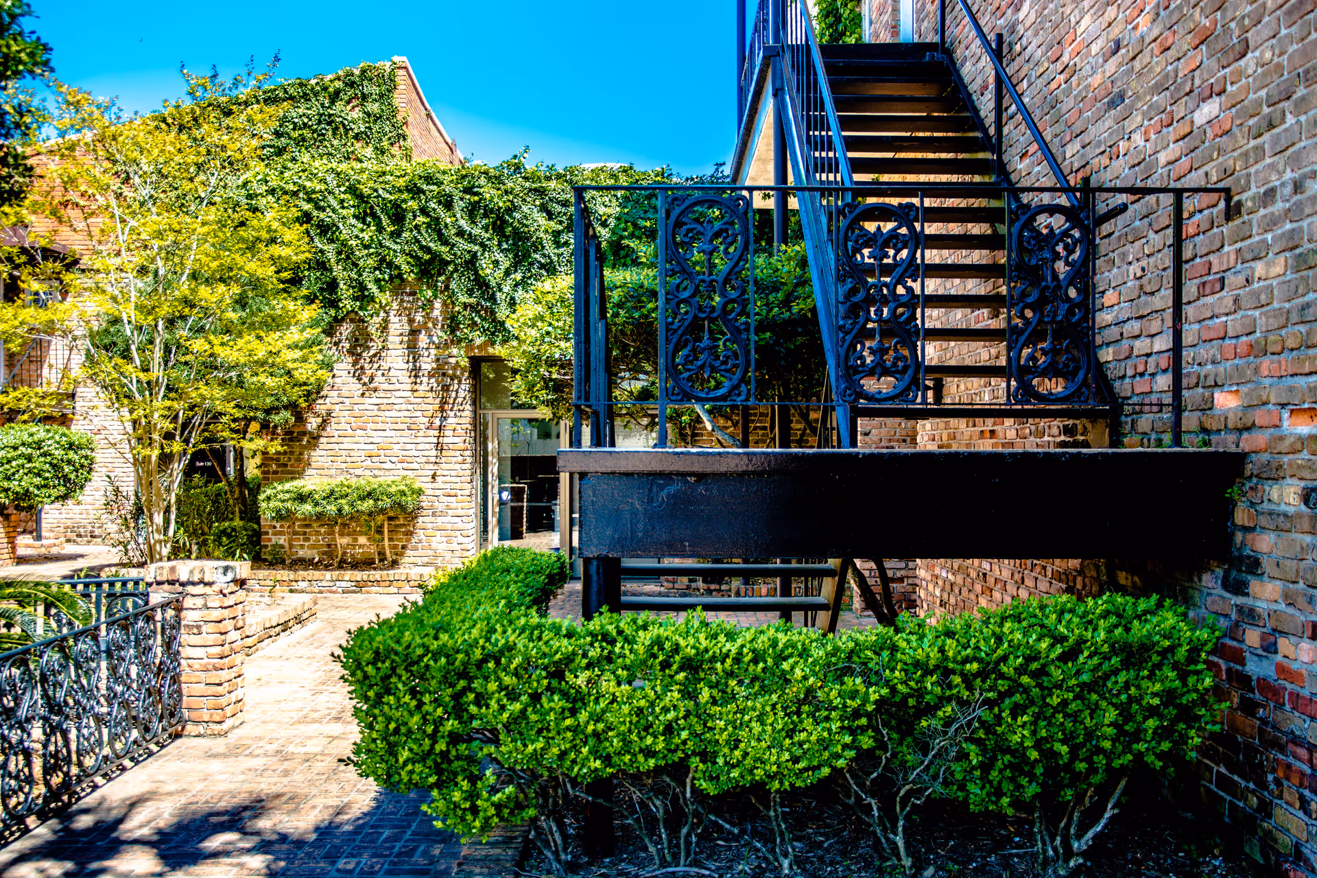 Brick building exterior with wrought-iron staircase, greenery, and hedges in a sunny courtyard.