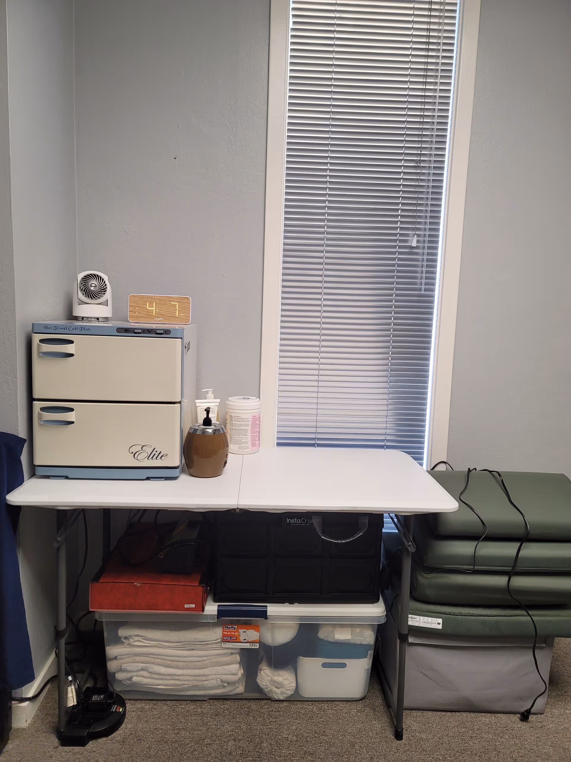 Treatment-room corner with a small cabinet, supplies, and a window with blinds.
