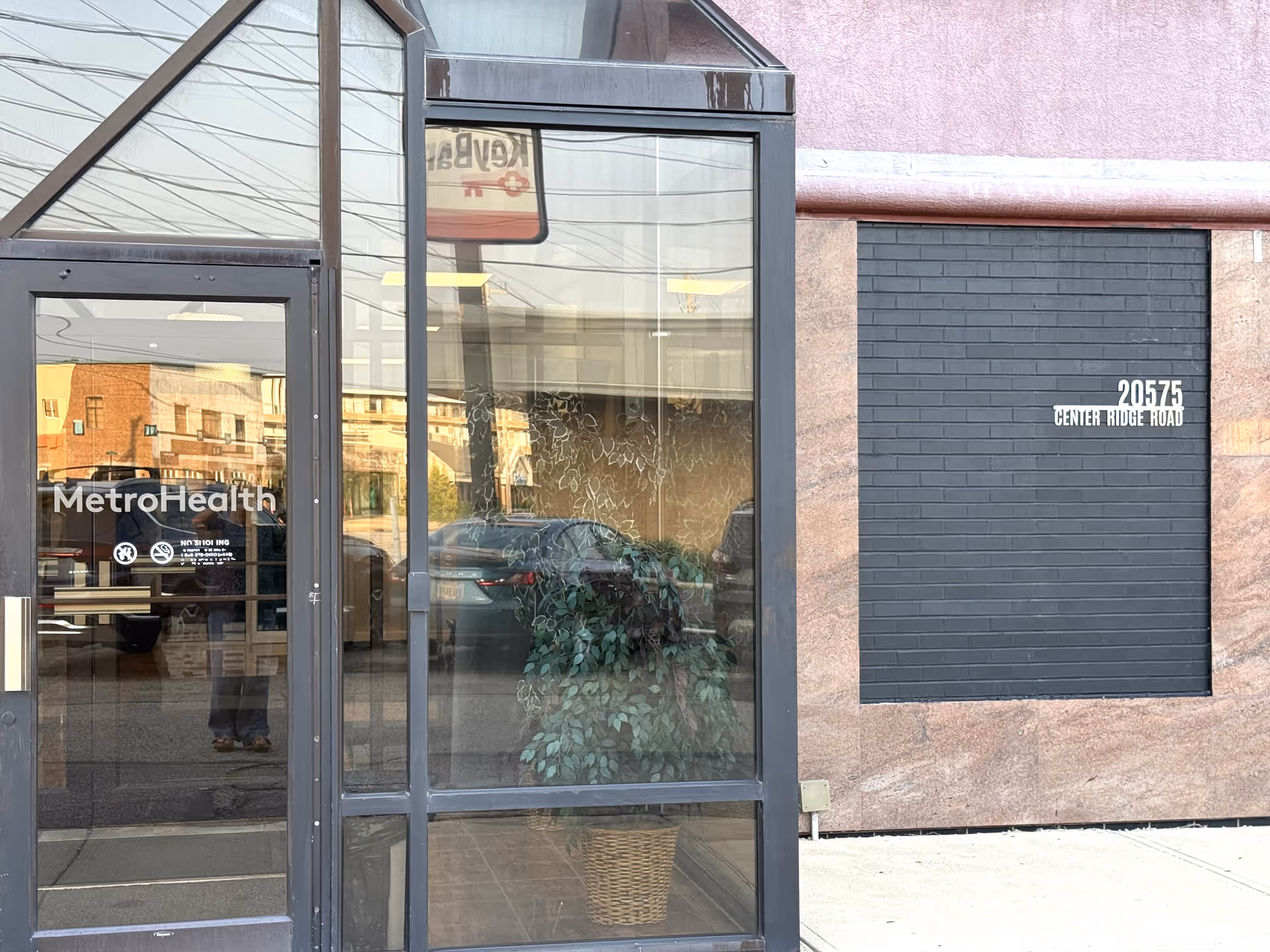 Glass-front building entrance with doors, signage, reflections, and a plant inside.