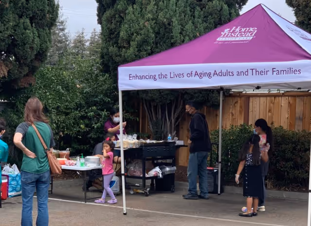 Outdoor outreach booth under a purple canopy with people gathered around tables