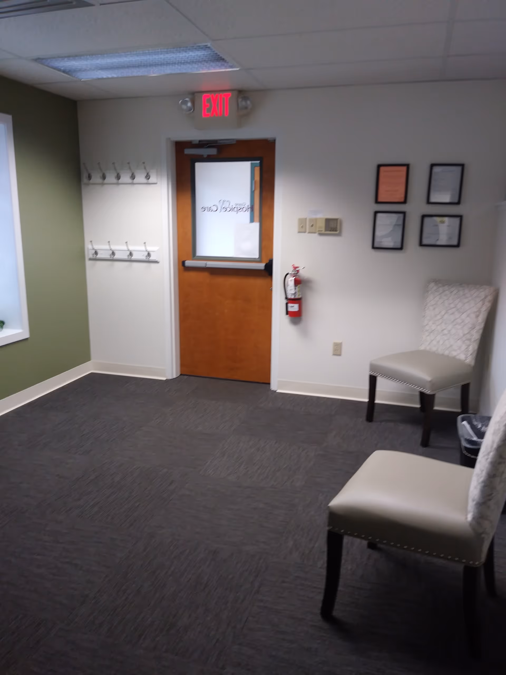 Interior hallway with exit sign, wooden door, chairs, coat hooks, and framed certificates.