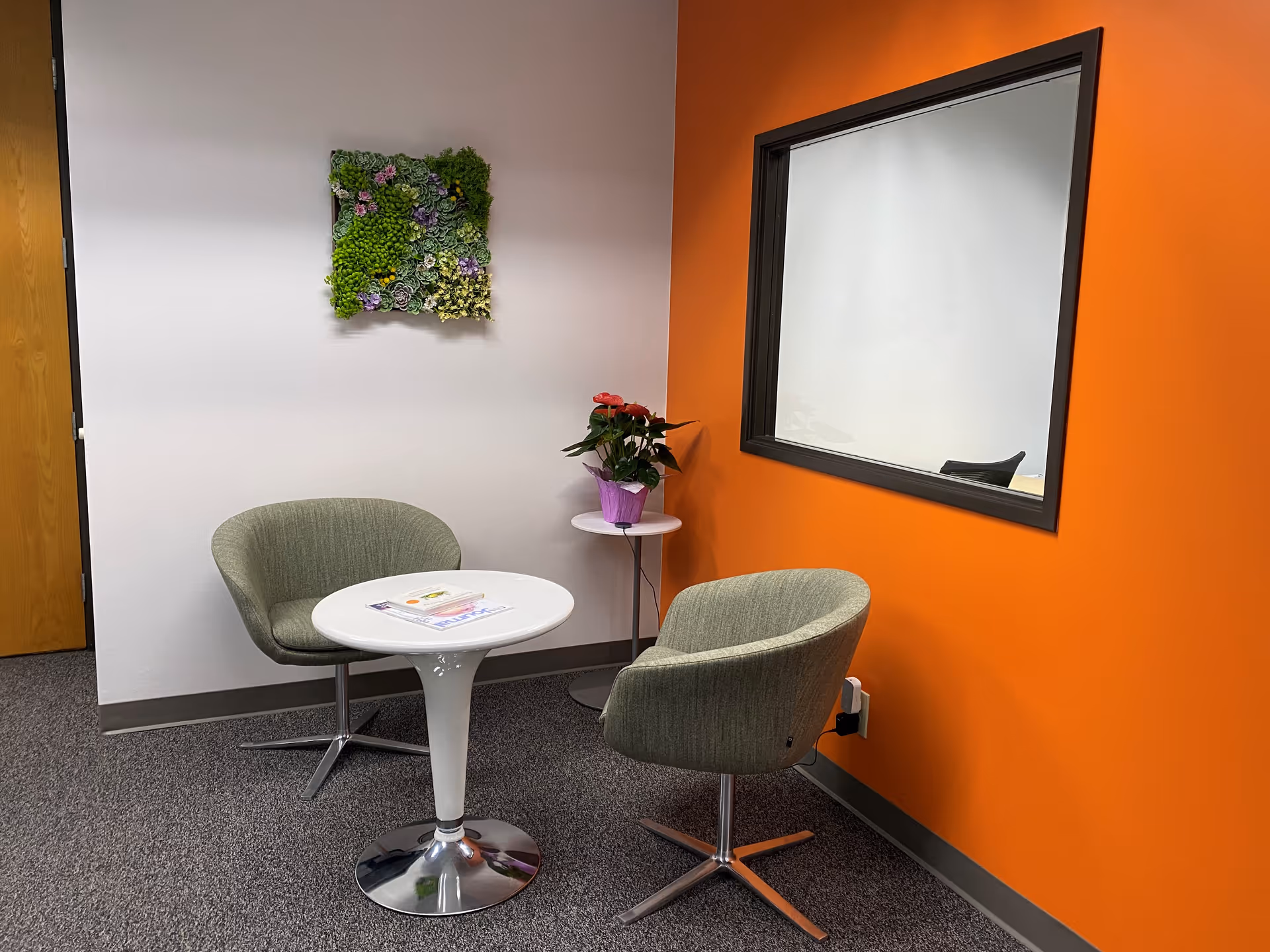 Two green chairs around a white round table in a bright orange-walled lounge.