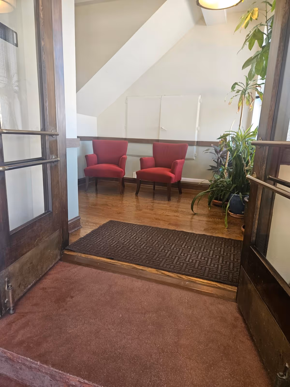 Indoor lobby area with two red chairs, a potted plant, and wooden entry doors.
