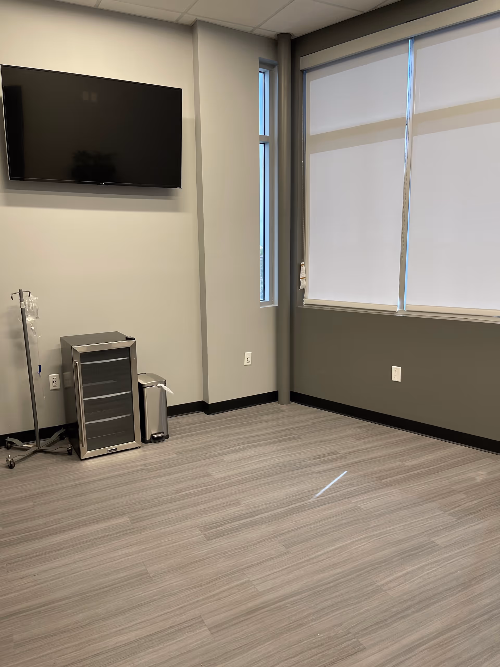 A clean, modern treatment room with a TV, IV pole, small cabinet, and window blinds.