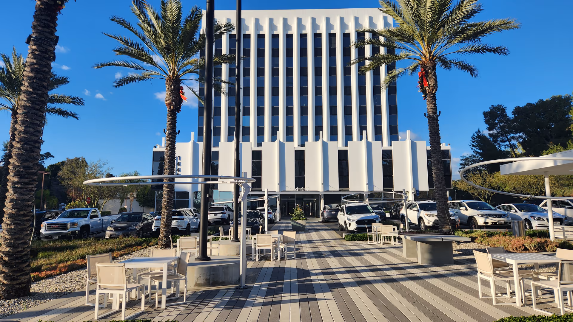 Modern multi-story building entrance framed by palm trees, with outdoor seating and parked cars.
