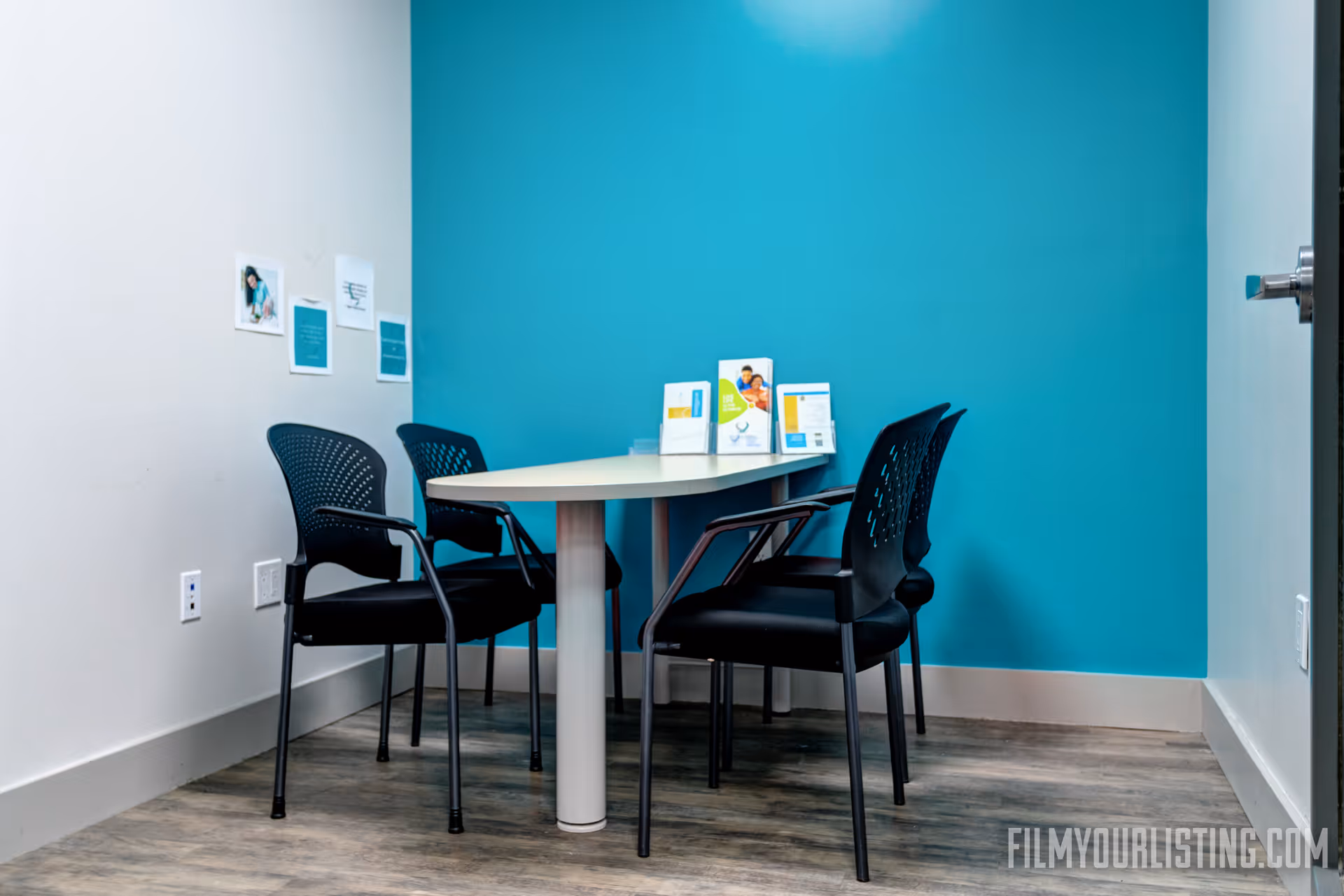 Small meeting room with a white table and four black chairs against teal walls.