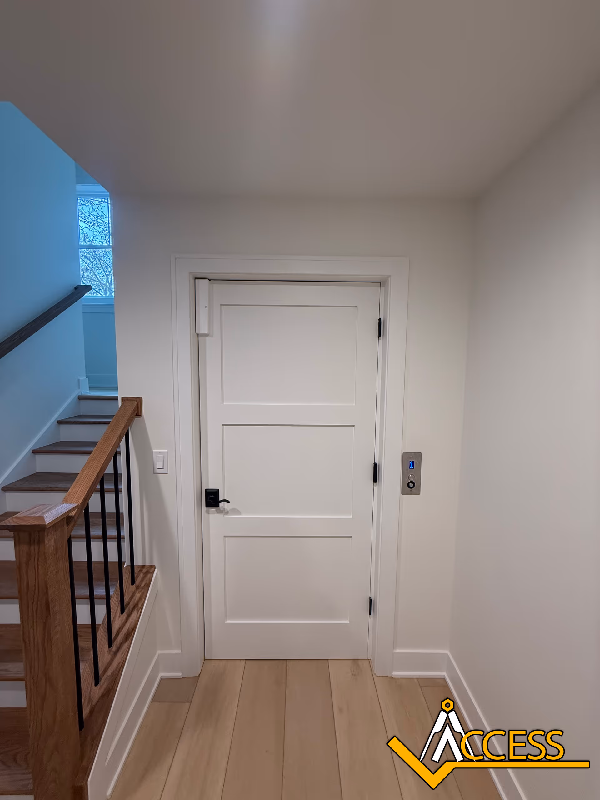White interior hallway with a closed door, wooden stairs, and an elevator panel.