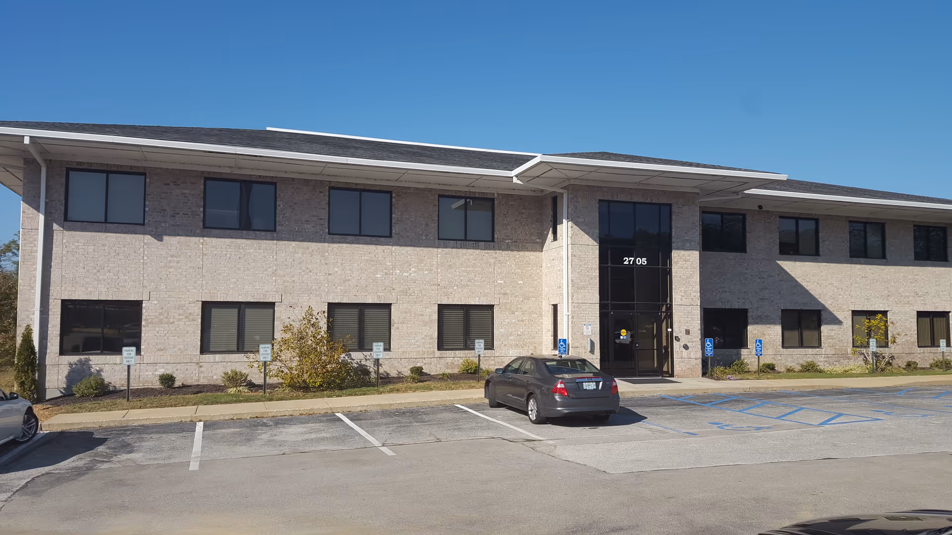 Two-story brick facility exterior with glass entrance, parked car, and accessible parking spaces nearby.