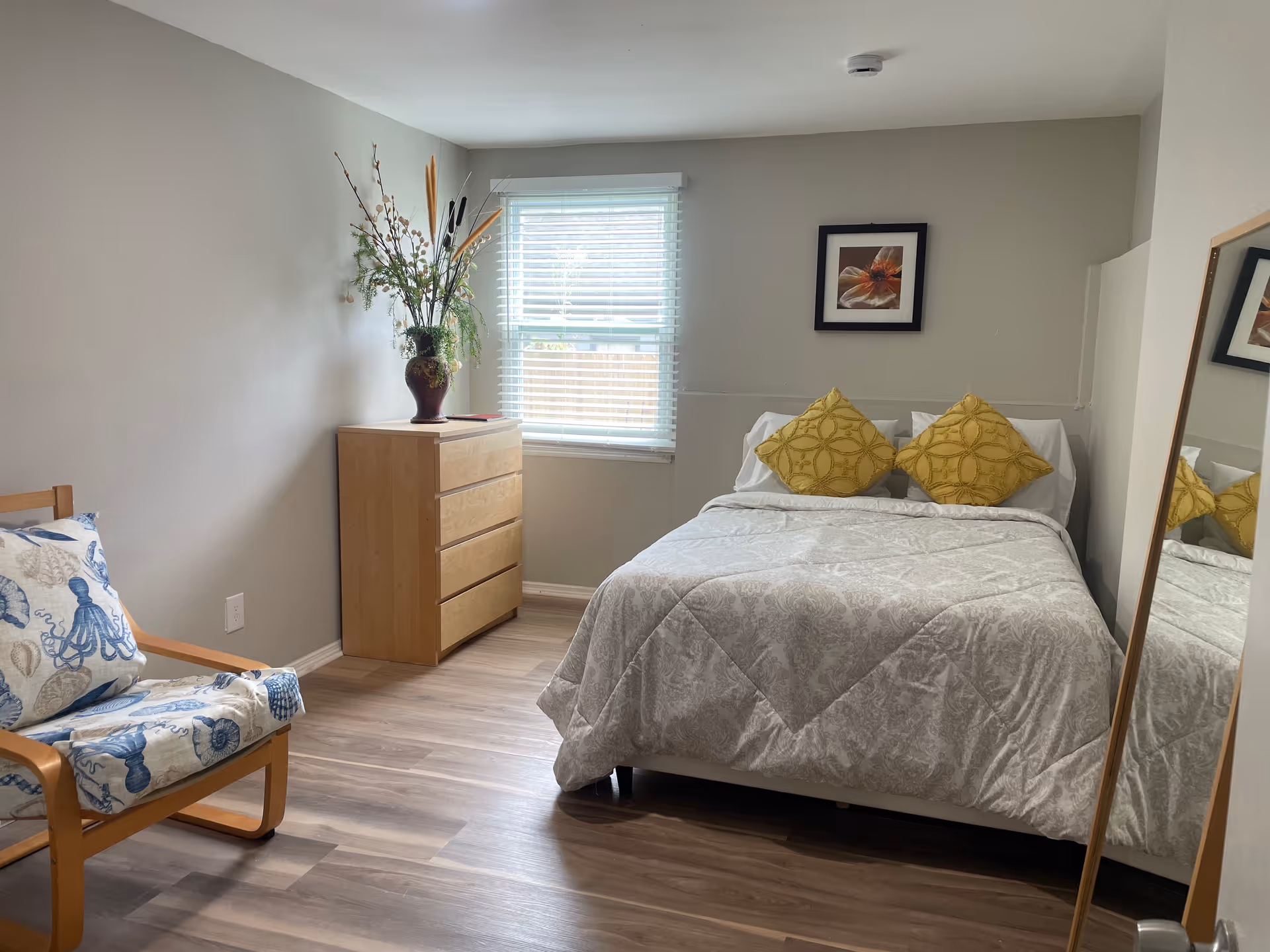 A tidy bedroom in a care home with a bed, dresser, chair, and mirror.