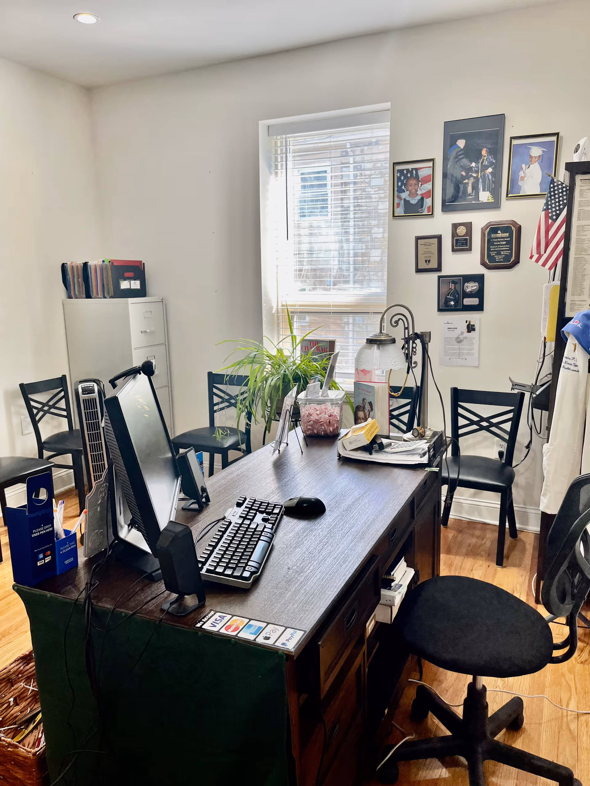 Office desk with computer, plant, wall plaques, and several chairs in a bright room.