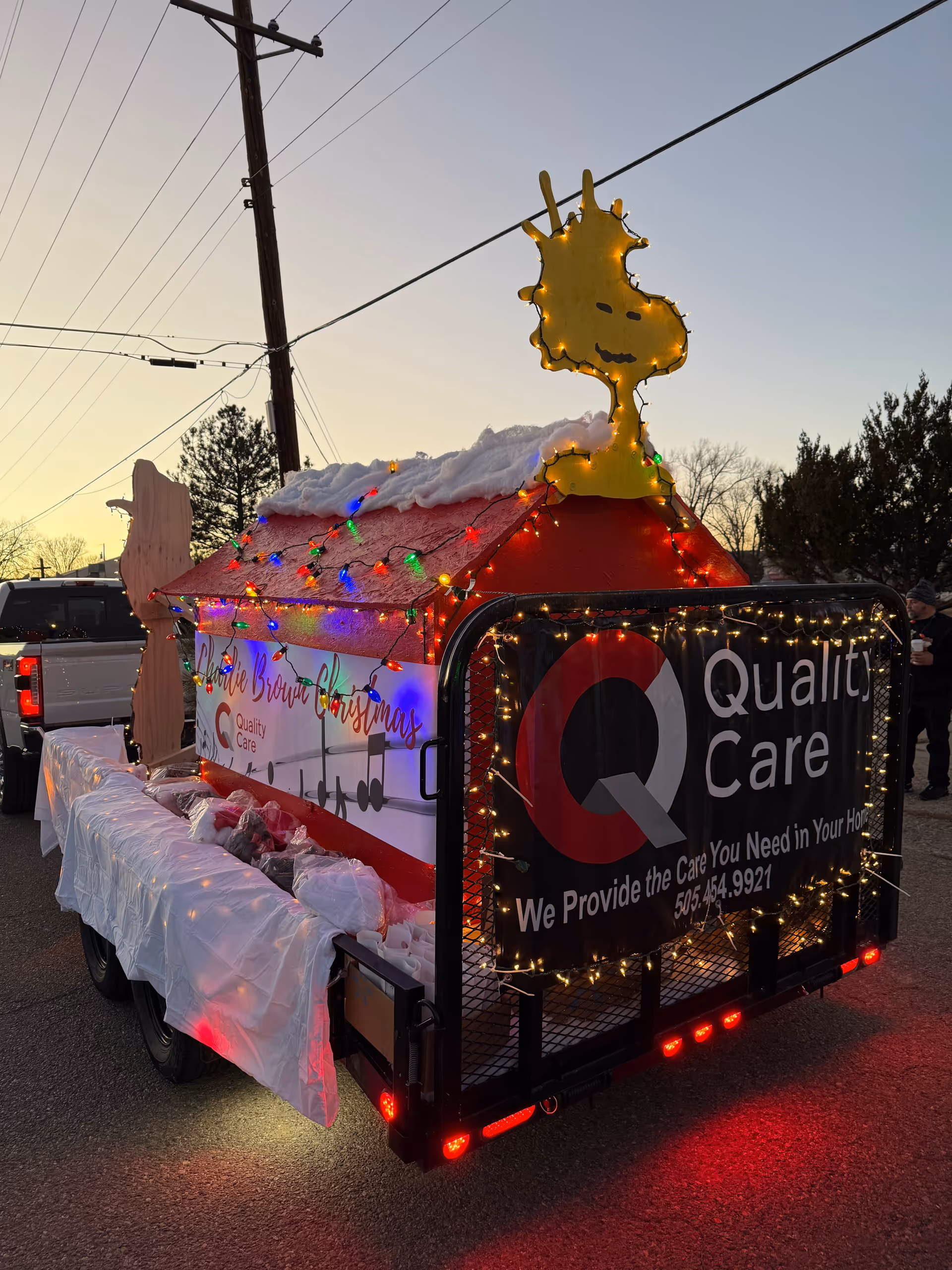Holiday-lit trailer float featuring a giraffe-shaped light and Quality Care banner at dusk.