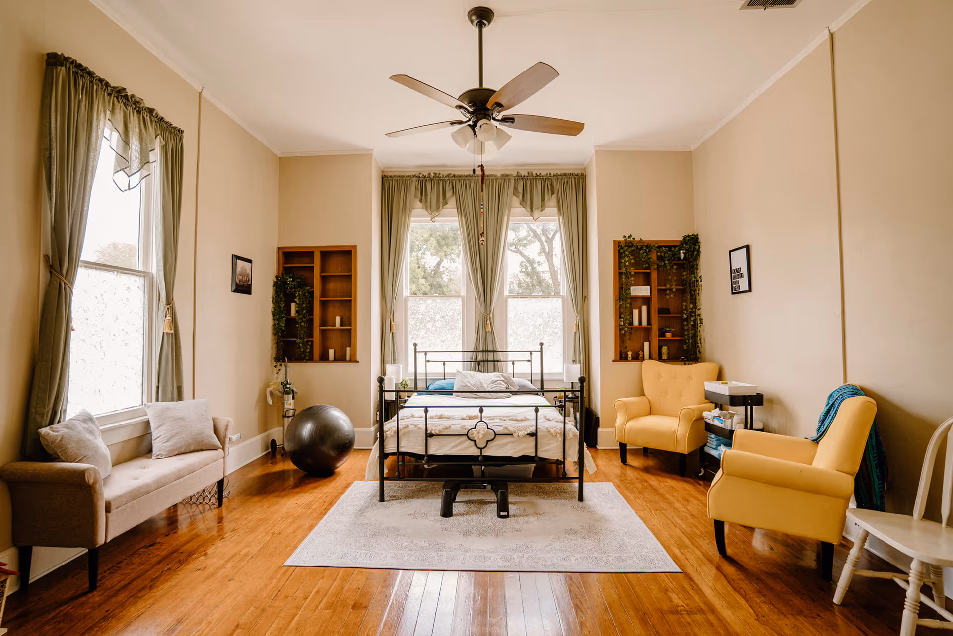 Bright bedroom with a metal bed, seating, and large front windows with green curtains.