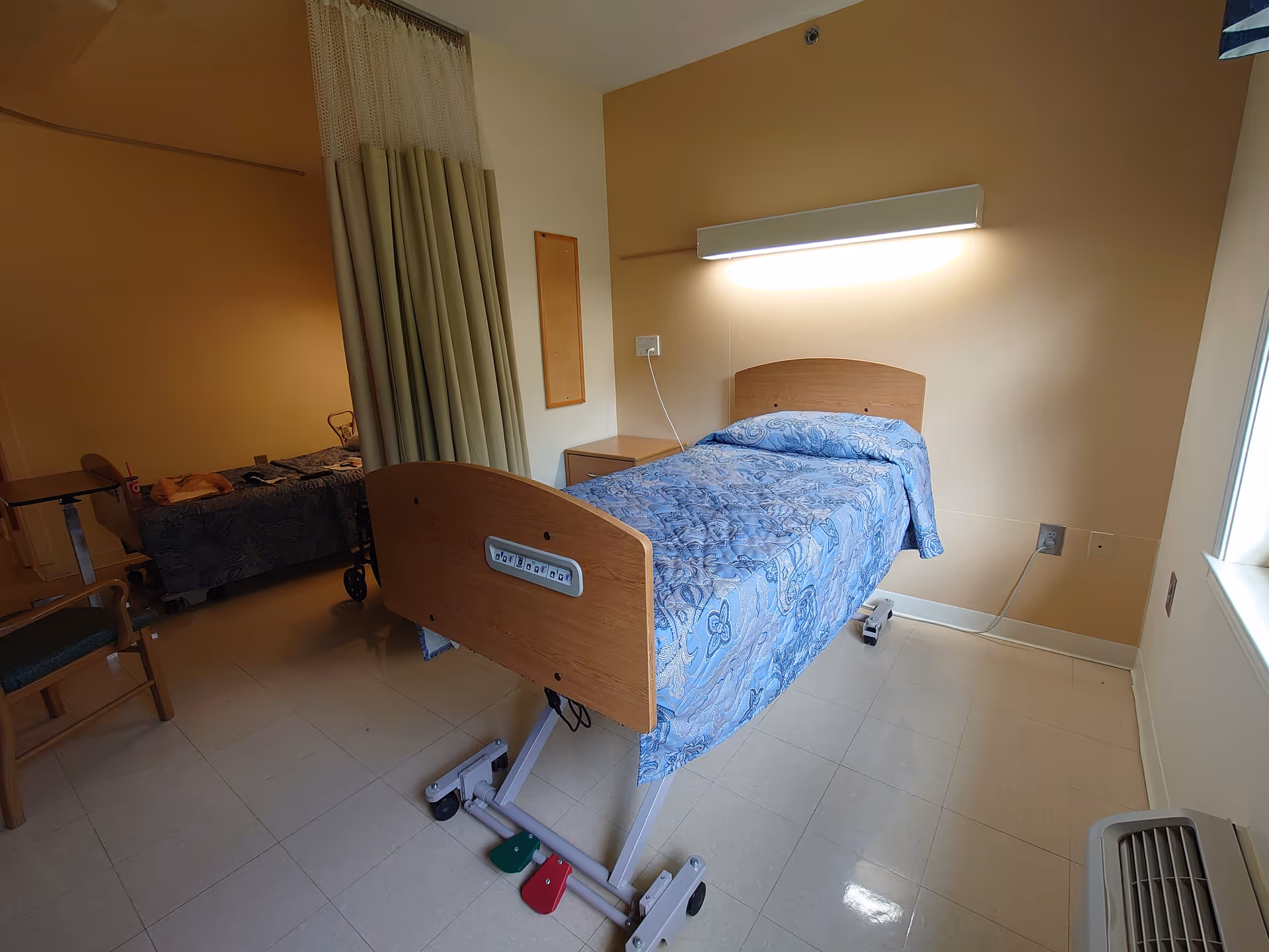 A hospital-style bedroom in a senior facility with a blue-patterned bed and partition curtain