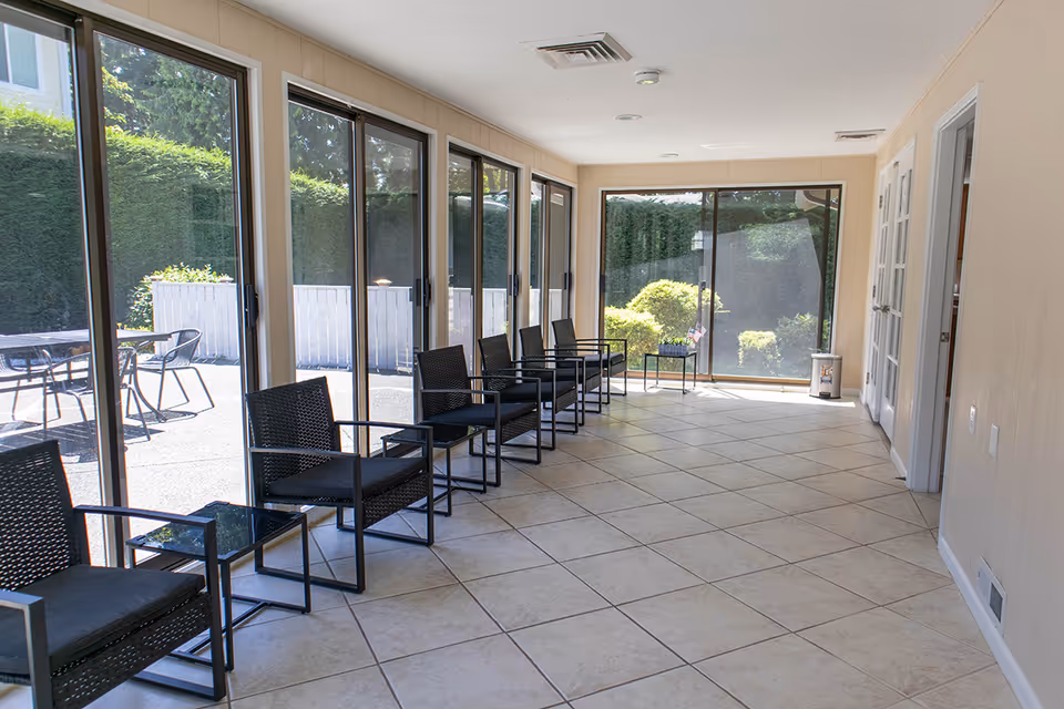 Long sunlit hallway with glass walls, row of black wicker chairs along tiled floor.