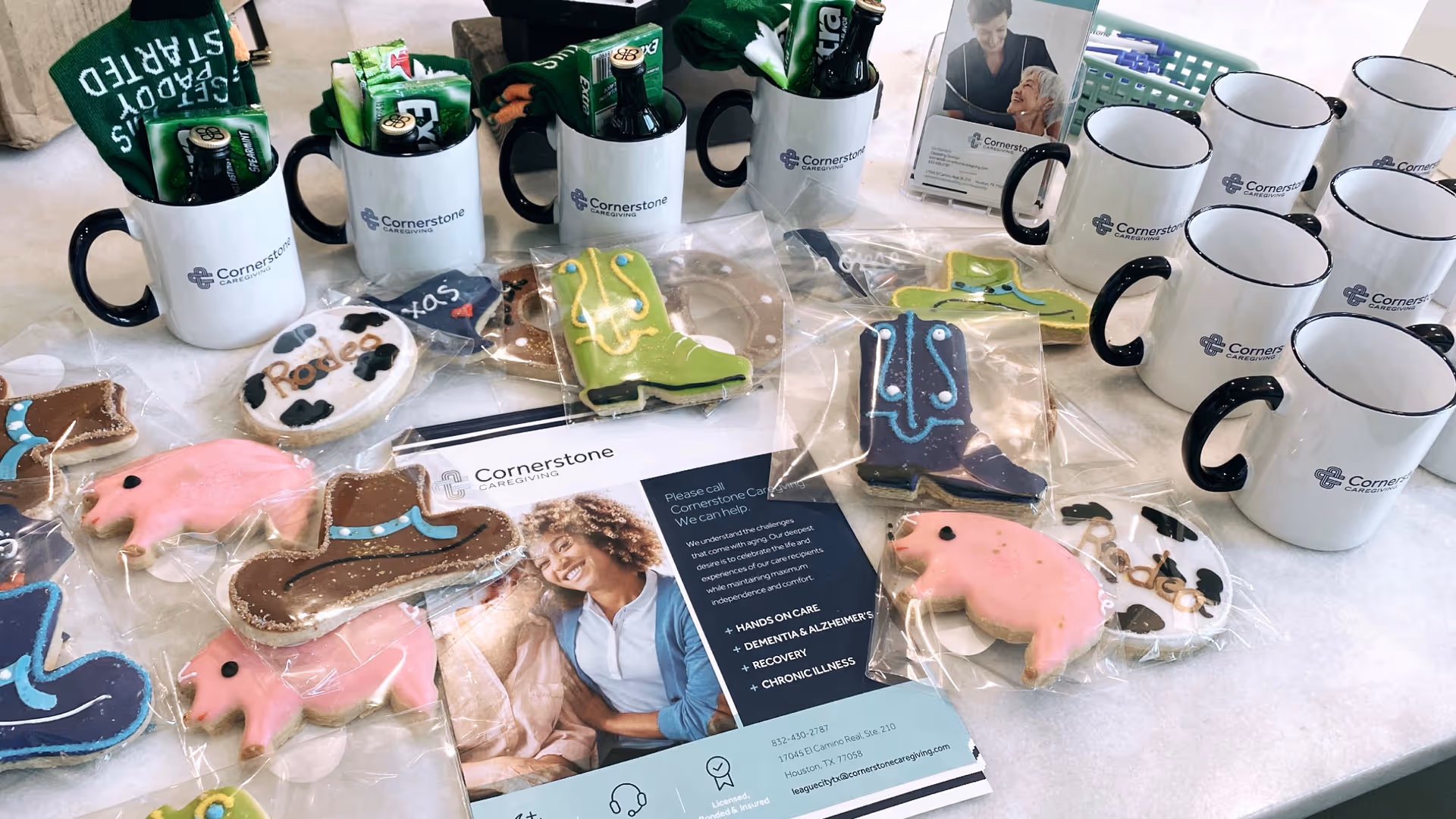 Branded Cornerstone Caregiving mugs, decorated cookies, and brochures on a table.