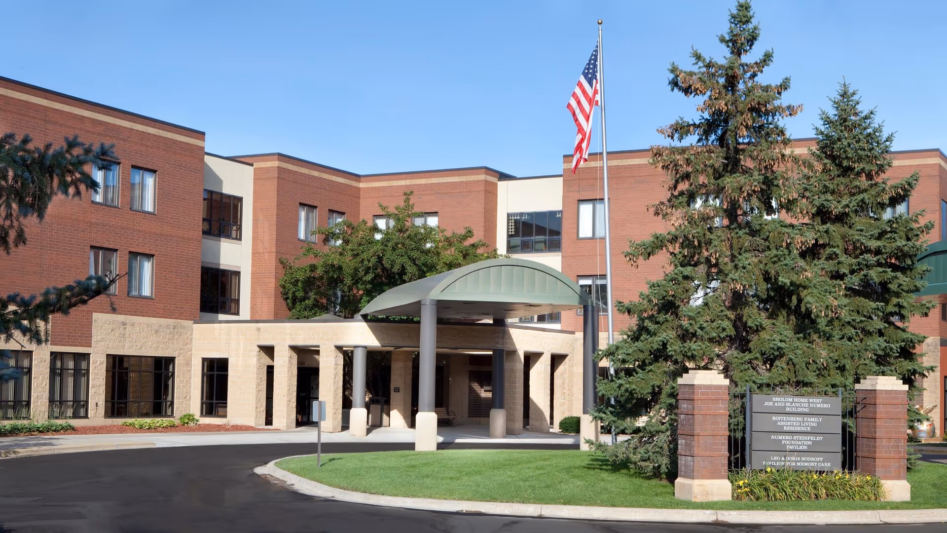 Exterior view of a brick senior living facility with entrance canopy, American flag, and trees.