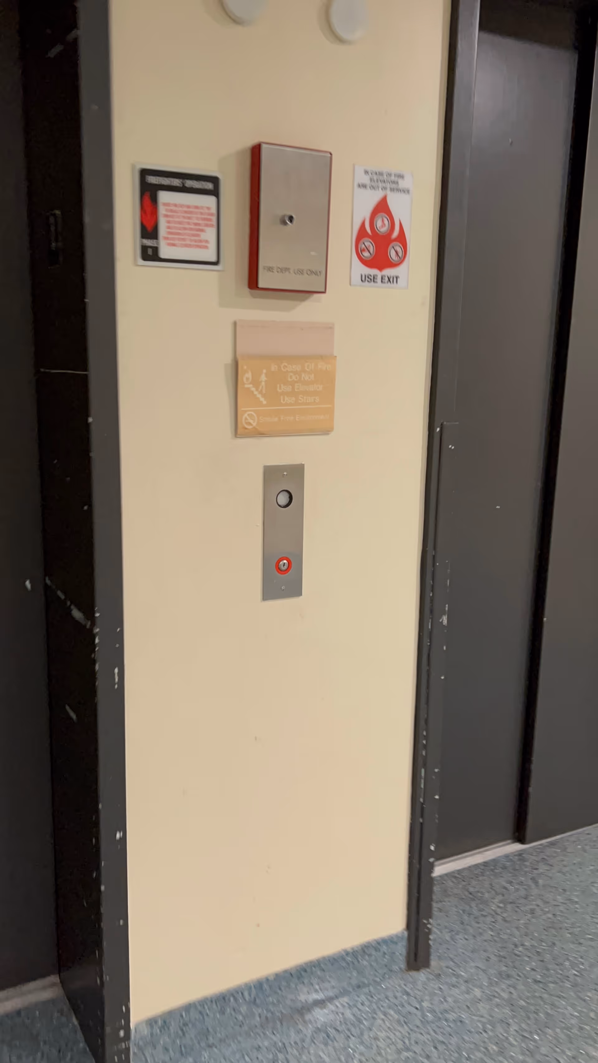 An elevator panel with buttons and exit signs on a beige hallway wall.