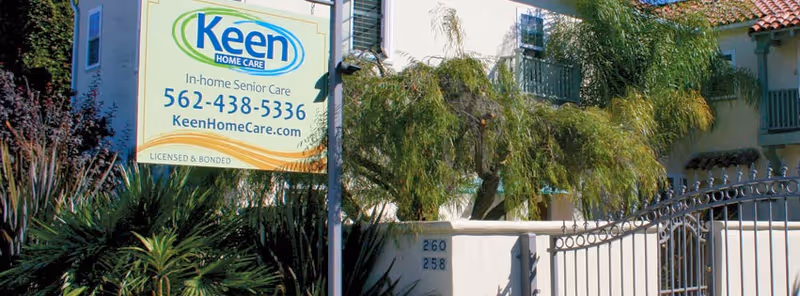 Keen Home Care sign at a gated building entrance with tropical plants.