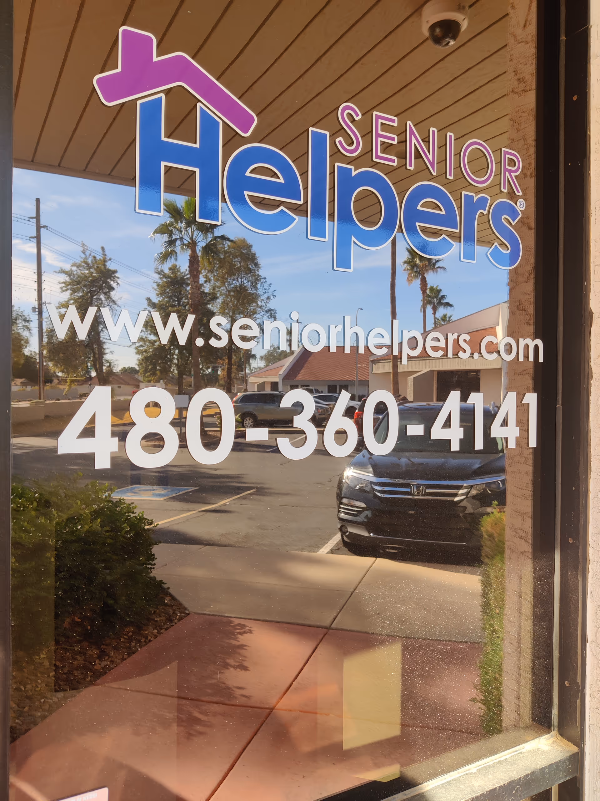 Glass storefront window shows Senior Helpers sign, website, phone, with parking lot reflections.