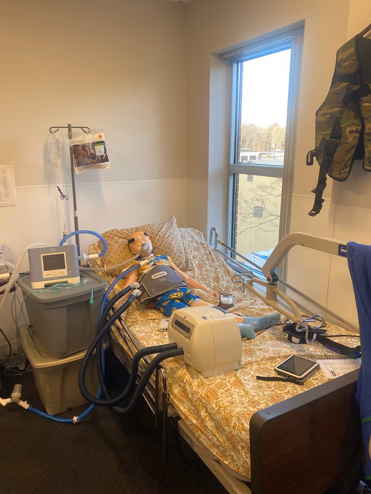 Hospital-style patient room with bed, monitors, ventilator equipment, and a window.