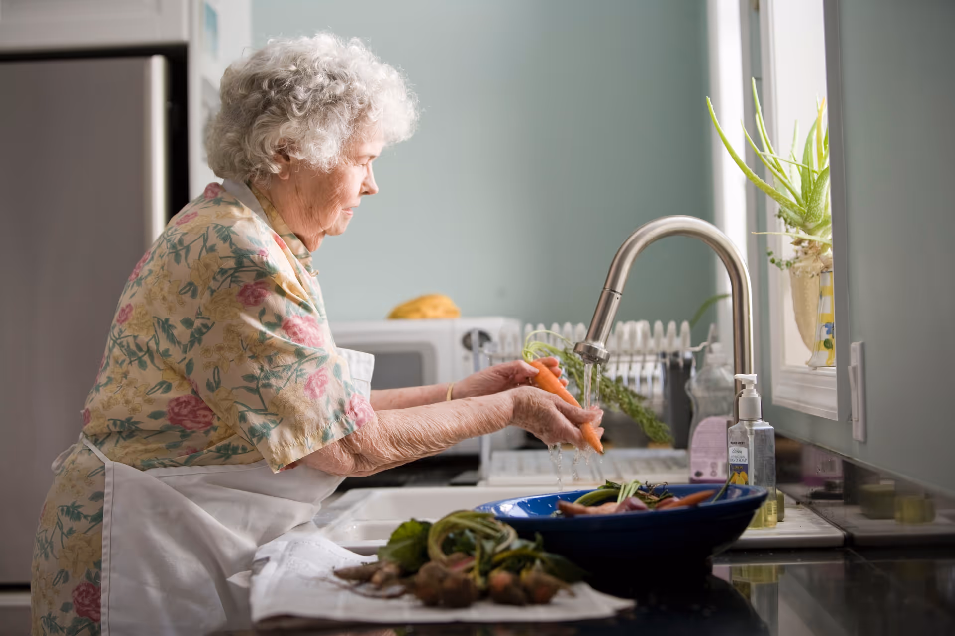 Elderly woman wearing a floral apron rinses vegetables at a kitchen sink.