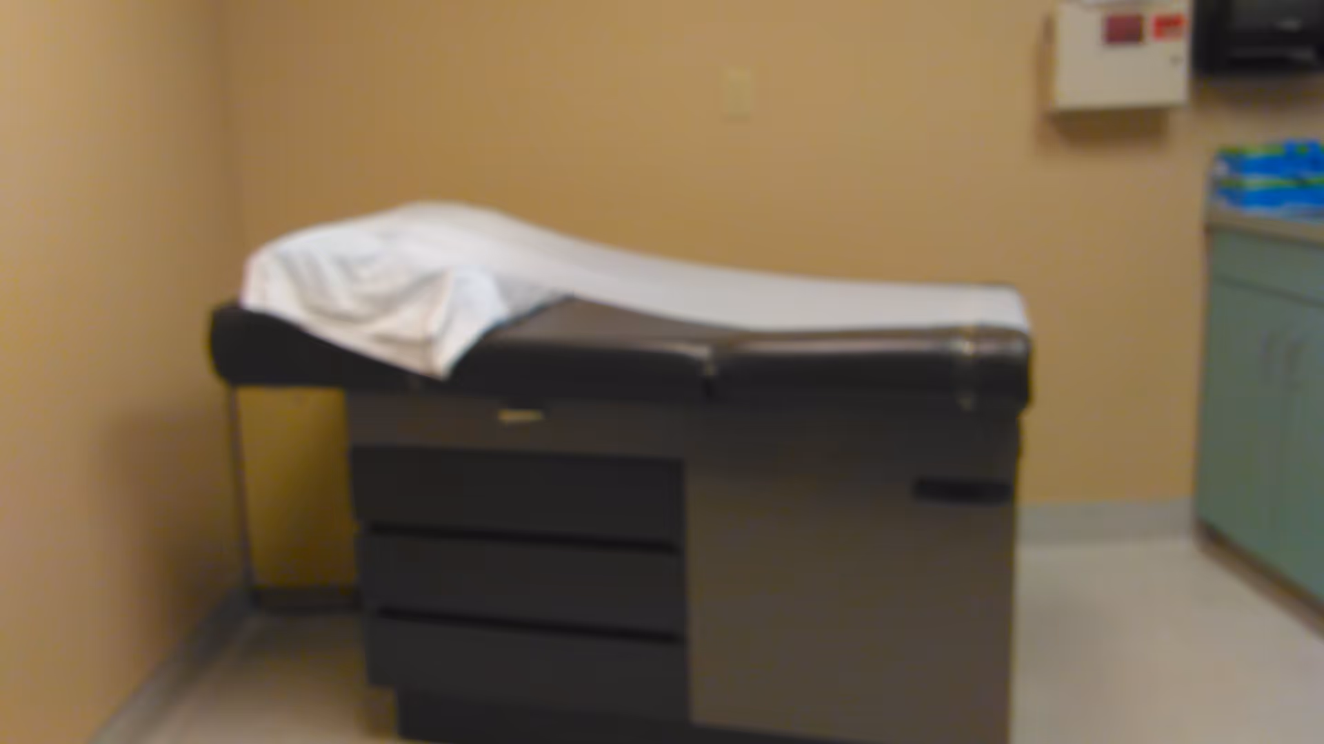 A medical examination table in a small clinic room with beige walls and green cabinets.